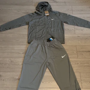 Nike grå tracksuit - Nike träningsset i grått med jacka och byxa. Jackan har huva, dragkedja och vita Nike-loggor på bröst och byxa. Lätt material, perfekt för löpning eller träning. Byxorna har resår i midjan och sidofickor. Snygg och sportig look.