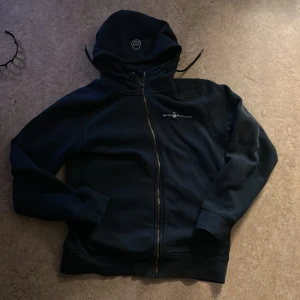 Svart hoodie från Sail Racing M - Svart hoodie från Sail Racing i storlek M med broderad logga på bröstet och huvan. Tröjan har hel dragkedja framtill, huva med snörning och ribbade muddar. Materialet är mjukt och känns som polyester, perfekt för kyliga dagar. Båda fickorna på sidorna är trasiga och lite på dragtjedjan