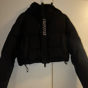 Pufferjacka Trapstar - Säljer en svart croppad pufferjacka från Trapstar med broderad logga i silver på ryggen och framtill. Jackan har huva, dragkedja och snörning i nederkant för extra street vibe. Perfekt för dig som vill sticka ut med en fet vinterjacka.