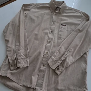 Beige skjorta Marlboro Classics M - Klassisk beige skjorta från Marlboro Classics i storlek medium. Skjortan har button-down krage, bröstficka med logotyp och knappar hela vägen. Tillverkad i mjuk bomull med långärmad passform och normal siluett. Perfekt för dig som gillar stilrena och tidlösa plagg.