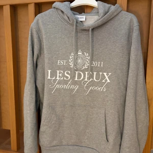Grå hoodie från Les Deux XS - Grå hoodie från Les Deux med vit tryckt logga och text på bröstet. Tröjan har huva med snörning, känguruficka och långa ärmar. Mjuk insida i bomullsmix som gör den extra skön. Perfekt för chill dagar och streetwear-stil.