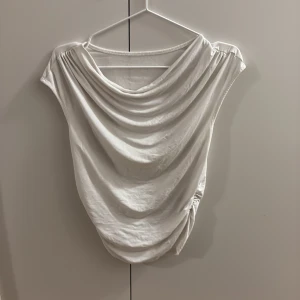 Vit draperad linnetopp - Säljer en vit linnetopp med snygg drapering framtill och lätt rynkade axlar. Toppen har en lös passform och är ärmlös, perfekt för dig som gillar stilrena och enkla plagg med lite extra detalj. Materialet känns mjukt och luftigt.