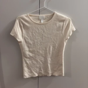 Beige ribbad topp från H&M - Säljer en beige ribbad topp från H&M i storlek M. Toppen har rund halsringning och korta ärmar, med en slim passform som sitter snyggt. Materialet är mjukt och stretchigt, perfekt för en clean och enkel look.
