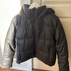 Matt Svart pufferjacka med huva - Svart pufferjacka med snygg huva och dragkedja framtill. Jackan har quiltad design och är tillverkad i ett skinnliknande material som ger en cool vibe. Perfekt för dig som gillar streetstyle och vill hålla dig varm under vintern. Dragsko vid huvan för extra stilpoäng.