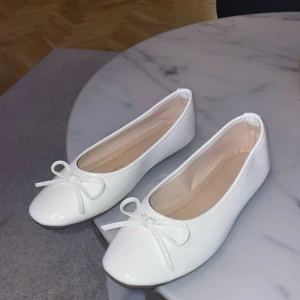 Vita ballerinaskor med rosett - Snygga vita ballerinaskor med rund tå och söt rosettdetalj framtill. Skorna har en platt sula och är gjorda i ett glansigt syntetmaterial som ger en fräsch och stilren look. Perfekta för dig som gillar klassisk och enkel stil. Helt oanvända! Säljs pga liten för mig