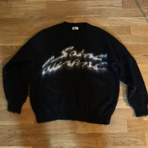 Saint Laurent svart stickad tröja - Svart stickad tröja från Saint Laurent Paris med vit fuzzy logga framtill. Oversized passform och långärmad modell, perfekt för dig som vill ha en statement piece med lyxig känsla. Materialet är mjukt och fluffigt, vilket ger en cool och avslappnad vibe. Storlek M 