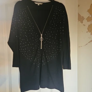 Svart tröja med dragkedja från Cellbes - Svart långärmad tröja från Cellbes med silvriga små nitar framtill och en snygg dragkedja med ringdetalj. Tröjan har en lös passform och är tillverkad i en mjuk mix av polyester, viskos och elastan. Perfekt för dig som gillar stilrena detaljer.