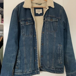 Blå jeansjacka med teddyfoder Pull&Bear - Säljer en blå jeansjacka från Pull&Bear i storlek L med mjukt vitt teddyfoder och krage. Klassisk denimlook med två bröstfickor, knappar framtill och detaljer i kontrastsömmar. Perfekt för kyligare dagar när du vill ha både stil och komfort.