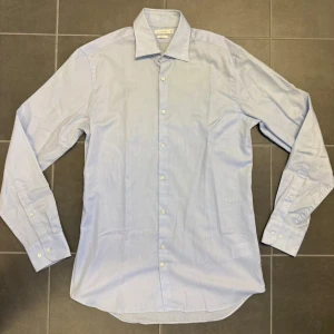 Ljusblå skjorta J.Lindeberg - Snygg ljusblå skjorta från J.Lindeberg i slim fit-modell. Skjortan har klassisk krage, långa ärmar och knäppning framtill. Tillverkad i bomull för en skön känsla och stilren look. Perfekt för dig som gillar en clean och modern stil.