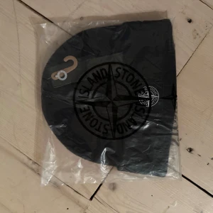 Svart mössa från Stone Island - Svart mössa från Stone Island med klassisk logotyp på framsidan. Snygg och enkel design som passar till de flesta stilar. Mössan är tillverkad i mjukt material och levereras i originalförpackning.