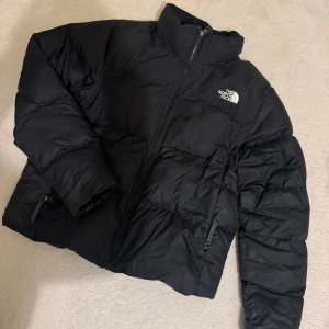 Svart pufferjacka från The North Face - Säljer en svart pufferjacka från The North Face med hög krage och dragkedja framtill. Jackan har två sidofickor med dragkedja och en klassisk logga på bröstet. Perfekt för kalla dagar och riktigt snygg till streetwear-stilen.