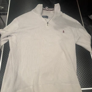Vit stickad polotröja Polo Ralph Lauren - Vit stickad polotröja från Polo Ralph Lauren med halv dragkedja och broderad logga i vinrött på bröstet. Tröjan har ribbade detaljer längs sidorna och muddar, samt hög krage med randig insida. Perfekt för lager-på-lager under kyliga dagar.