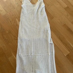 Vit stickad långklänning med snörning - Vit stickad långklänning med snyggt hålmönster och djup v-ringning med snörning framtill. Klänningen har slits på sidan och är ärmlös. Materialet är mjukt och luftigt stickat.