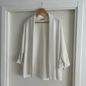 Vit blus kofta - Stilren vit blus kofta med omlottkänsla och lös passform. Jackan har långa ärmar och ett mjukt, lätt tyg som faller snyggt. Perfekt för lager-på-lager och enkel att matcha med olika outfits.