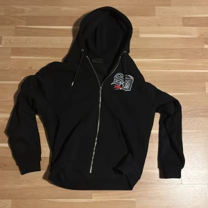 Supply & demand zip hoodie - Svart hoodie med huva och snörning från supply & demand, hel dragkedja framtill och ett coolt tryck på bröstet i vitt, grått och rött. Tröjan har långa ärmar och ribbade muddar. Perfekt för en avslappnad stil.