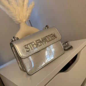 Silver axelväska från Steve Madden - Snygg axelväska i metallic silver från Steve Madden med croco-mönstrad yta och kedjerem. Väskan har ett stort logotyp-spänne framtill och stilren, boxig form. Perfekt för dig som vill sticka ut med accessoarerna. Aldrig använd och i toppskick! 