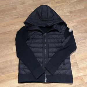 Moncler cardigan - Svart dunjacka från Moncler med quiltad front och huva. Jackan har ribbstickade ärmar och sidor, dragkedja framtill och Moncler-logga på ärmen. Perfekt för kyliga dagar med sin kombination av dun och stickat material.