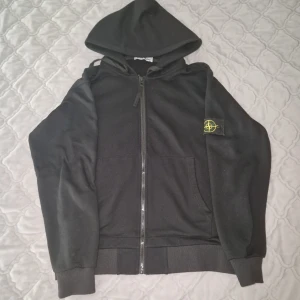 Svart hoodie från Stone Island - Svart hoodie från Stone Island med dragkedja framtill och klassisk logotyp på vänster ärm. Tröjan har huva, känguruficka och ribbade muddar. Perfekt för dig som gillar streetwear och vill ha en stilren look.