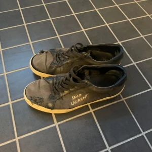 Svarta Saint Laurent sneakers - Svarta sneakers från Saint Laurent med vit sula och handskriven logga på sidan. Skorna har snörning, rund tå och är tillverkade i en mix av skinn och textil. Klassisk låg modell med slitna detaljer för en edgy look.