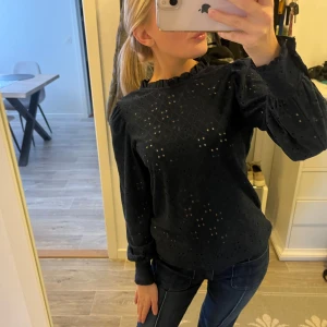 Mörkblå blus med broderade detaljer - Säljer en mörkblå blus med broderade hålmönster och volangkant vid halsen. Blusen har långa ärmar med lätt puff och resår vid ärmslut.