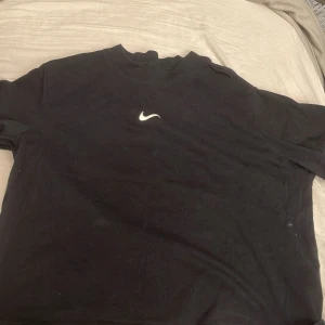 Svart Nike t-shirt med logga - Svart t-shirt från Nike med klassisk vit swoosh-logga broderad på bröstet. Enkel och stilren design med rund hals och korta ärmar. Perfekt för en sportig och avslappnad look.