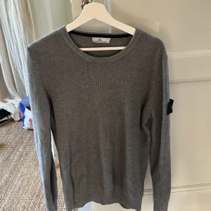Grå stickad tröja från Stone Island - Säljer en grå stickad tröja från Stone Island med klassisk rund hals och ribbad struktur. Tröjan har lång ärm och den ikoniska Stone Island-loggan på vänster ärm. Perfekt för dig som gillar stilren och enkel design.