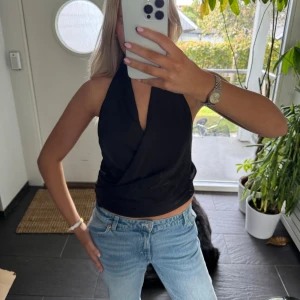 Svart omlott halterneck topp - Säljer en svart omlott halterneck topp med djup v-ringning. Toppen är ärmlös och har en croppad passform som sitter snyggt till jeans eller kjol. Perfekt för dig som gillar stilrena och trendiga plagg med en twist.