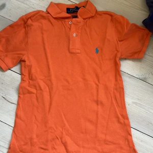 Orange pikétröja från Ralph Lauren, strl 8 - Snygg orange pikétröja från Ralph Lauren med klassisk krage och två knappar framtill. 
