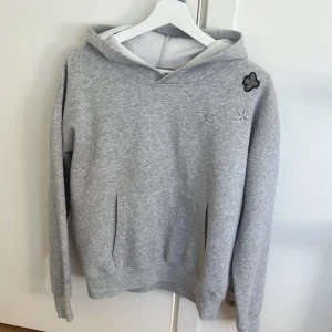 Grå hoodie från Hunkydory med brodyr - Gråmelerad hoodie från Hunkydory med broderade stjärnor och en liten blomma på bröstet. Tröjan har huva, magficka och är tillverkad i en mjuk mix av bomull och polyester. Perfekt för chill dagar och enkel att matcha med jeans eller leggings.