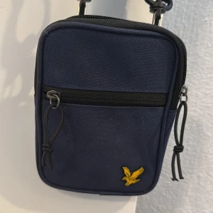 Mörkblå axelväska från Lyle & Scott - Mörkblå liten axelväska från Lyle & Scott med justerbar, avtagbar rem och gul broderad logga framtill. Väskan har ett huvudfack och en mindre ficka med dragkedja på framsidan. Perfekt storlek för mobil, plånbok och nycklar. 