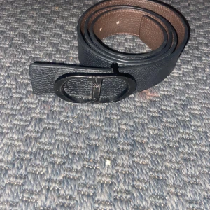 Svart och brunt skärp i skinn,Dio r - Stilrent bälte i svart och brunt skinn med snygg, oval metallspänne i svart. Bältet har en lätt texturerad yta och är vändbart med svart på ena sidan och brunt på andra. Perfekt för att lyfta vilken outfit som helst.