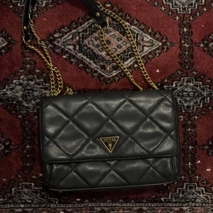 Svart quiltad axelväska från Guess - Snygg svart axelväska från Guess med quiltad design och guldfärgade kedjor. Klassisk rektangulär form med lock och ikonisk Guess-logga framtill. Väskan är tillverkad i skinnimitation och har en lyxig känsla som lyfter varje outfit. Några repor på loggan därav priset❤️