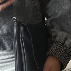 Svart mocka och skinn axelväska - Stilren svart axelväska. Väskan har ett avtagbart axelband och enkel, rektangulär form. Perfekt för dig som gillar minimalistisk design och vill ha en väska som sticker ut med sina olika material.