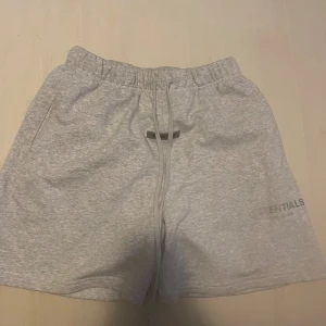 Grå Essentials Fear of God shorts - Säljer ett par ljusgrå shorts från Essentials Fear of God. De har elastisk midja med snörning, två sidofickor och en bakficka. Mjuk bomullskvalitet och logga tryckt på vänster ben. Perfekt för chill dagar och streetwear vibes.