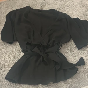 Svart omlottblus med knyt från SHEIN - Svart omlottblus från SHEIN i storlek XS. Blusen har korta ärmar, v-ringning och ett brett knytband i midjan som ger en snygg siluett. Tillverkad i ett lätt och flowy material som känns luftigt och skönt. Perfekt för dig som gillar stilrena och trendiga plagg.