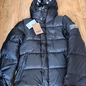 Svart pufferjacka från Burberry S - Svart pufferjacka från Burberry med huva och klassiskt rutigt foder. Jackan har dragkedja, knappar och flera fickor. Märkeslogga på ärmen och snygga detaljer. Perfekt för kalla dagar och riktigt stilren look. Kan ändras till en väst och påse medföljer.mer bilder kan skickas dm