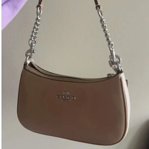 Beige axelväska från Coach i skinn - Snygg beige axelväska från Coach i äkta skinn med stilren form och silver- eller guldfärgade metalldetaljer. Väskan har dragkedja upptill, justerbar axelrem och diskret logga framtill. Perfekt storlek för det viktigaste och ger en clean look till din outfit.