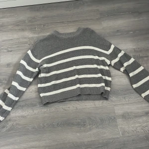 Gråvit randig stickad tröja - Säljer en ribbstickad tröja i grått och vitt med breda horisontella ränder. Tröjan har en croppad passform, rund halsringning och långa ärmar. Perfekt för dig som gillar en chill och stilren look.