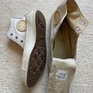 Vita Converse All Star i skinn - Säljer ett par vita Converse All Star high-top sneakers i skinn med klassisk rund tå och gummisula. Skorna har ikonisk Converse-logga på sidan och metallfärgade öljetter för snörning. Insidan är fodrad med textil för extra komfort. Perfekta för en clean och stilren look. 