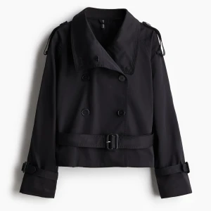 Svart trenchcoat med bälte - Stilren svart croppad trenchcoat med bred krage, dubbelknäppning och bälte i midjan. Jackan har långa ärmar med spännen vid ärmslut och axelklaffar för extra edge. Perfekt för dig som vill ha en klassisk look