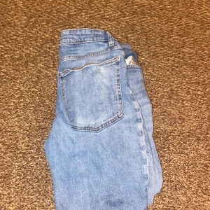 Ljusblå jeans från H&M, storlek 40 - Ljusblå jeans från H&M i klassisk femficksmodell med knäppning fram. Jeansen har raka ben och hög midja, tillverkade i bomull med snygga kontrastsömmar. Perfekta för en avslappnad och trendig look.