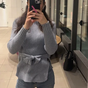 Ljusgrå ribbad stickad tröja med knyt - Superfin ljusgrå stickad tröja med ribbat mönster och knytband i midjan. Långärmad och mjuk, perfekt för kyligare dagar. Snygg till både jeans och kjol, enkel att styla för en trendig look.