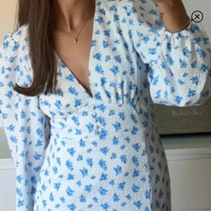 Blommig långklänning med puffärm - Långklänning i vit bas med blått blommönster, djup v-ringning och puffiga långärmar. Klänningen har knappar framtill och markerad midja för en feminin siluett. Perfekt för sommarens alla tillfällen.