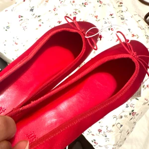 Röda ballerinaskor med rosett - Snygga röda ballerinaskor i skinn med rund tå och söt rosettdetalj framtill. Perfekta för dig som vill ha en poppig färg på fötterna och gillar klassisk, feminin stil.