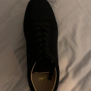 Christian Louboutin svarta sneakers - Säljer ett par svarta sneakers från Christian Louboutin med klassisk röd sula och guldfärgad logga på innersulan. Skorna har snörning och är tillverkade i ett tygmaterial. Perfekt för dig som vill sticka ut med exklusiva detaljer och ikonisk design.