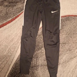 Svarta Nike träningsbyxor S - Svarta träningsbyxor från Nike med vit logga på låret och vita sidopaneler. Byxorna har en slim passform och är gjorda i mjukt syntetmaterial som andas. Perfekta för sport eller chill, med elastisk midja för skön komfort.
