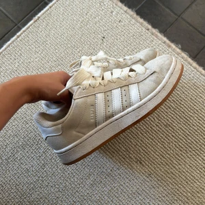 Adidas Campus sneakers i beige  - Säljer ett par Adidas Campus sneakers i beige med klassiska vita ränder på sidorna och gummisula. Skorna har rund tå, breda vita snören och Adidas-logga på hälen. Perfekta för dig som gillar stilrena och tidlösa sneakers.