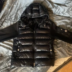 Svart Moncler Maya cardigan pufferjacka med huva - Säljer en svart cardigan från Moncler med glansig front, stickade ärmar och rygg med en väldigt skön och bekväm textur som håller värmen året runt. Jackan har en stor vadderad huva, dragkedja framtill och fickor med tryckknappar. Klassisk Moncler-logga på ärmen med nfc tagg som går att scanna. Fet mix av material för en modern och stilig look.