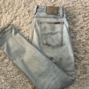 Ljusblå jeans från Nudie Jeans, W29 L30 - Säljer ett par ljusblå jeans från Nudie Jeans med klassisk vågsöm på bakfickan och orangea kontrastsömmar. Modellen har normal passform och raka ben. Tillverkade i mjuk bomullsdenim, perfekta för en avslappnad stil.