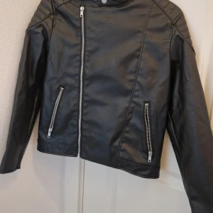Svart skinnjacka från H&M, stl 140 - Snygg svart skinnjacka från H&M i storlek 140. Jackan har coola detaljer med sned dragkedja, två fickor med dragkedjor och quiltade axelpartier. Perfekt för dig som vill ha en stilren och edgy look.